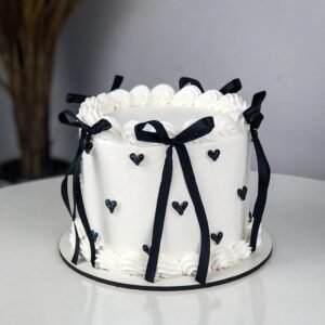 White frosting birthday cake with black satin ribbons and bows on a marble table with balloons, customized cake Karachi