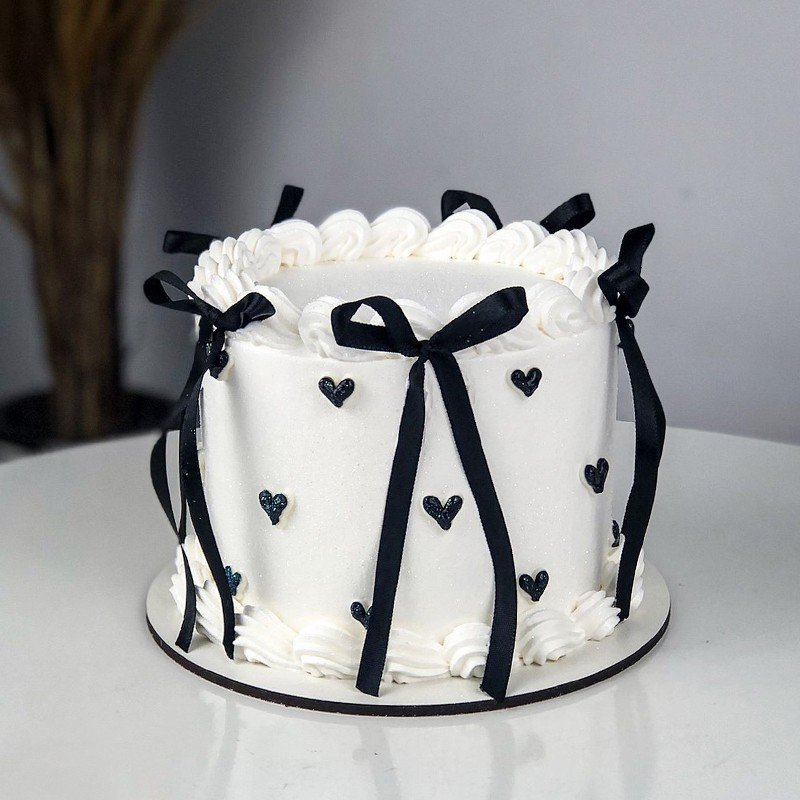White frosting birthday cake with black satin ribbons and bows on a marble table with balloons, customized cake Karachi